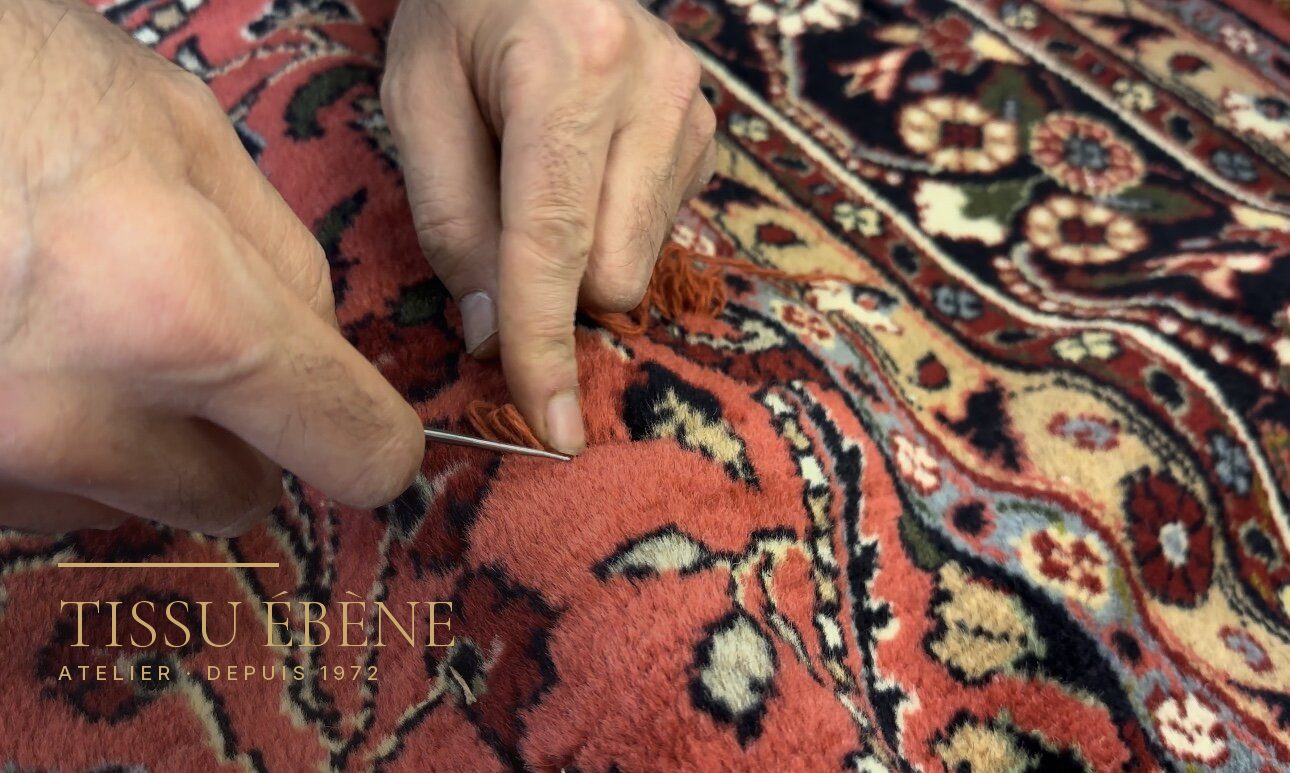 Mains tenant une aiguille et un brin de laine orangée sur un tapis noué rouge à fleurs